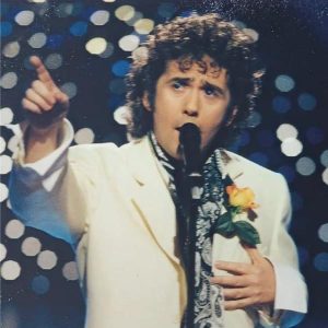 Christopher Nott as David Essex wearing white jacket and yellow rose button hole, singing in front of microphone, left hand over chest and right hand pointing forward