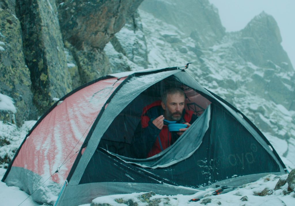 Thomas Salvador as Pierre in The Mountain / La Montagne , Pierre is sat in a dome tent, door partially unzipped eating a meal, the ten is set on a mountain surrounded by snow.