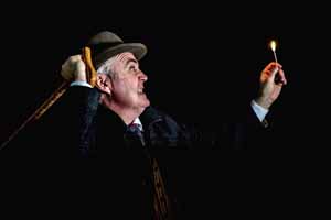 WATSON: The Final Problem publicity image, side ways pose of the Watson wearing a dark suit and hat, with right hand holding the hat and the left hand raised