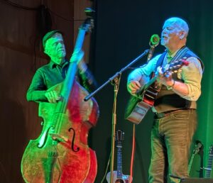 Mike Seal playing Bass and Joe Bayliss playing the guitar & singing into a microphone. Both men are standing.