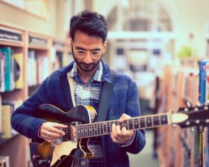 Amit Dattani playing a guitar standing in a library