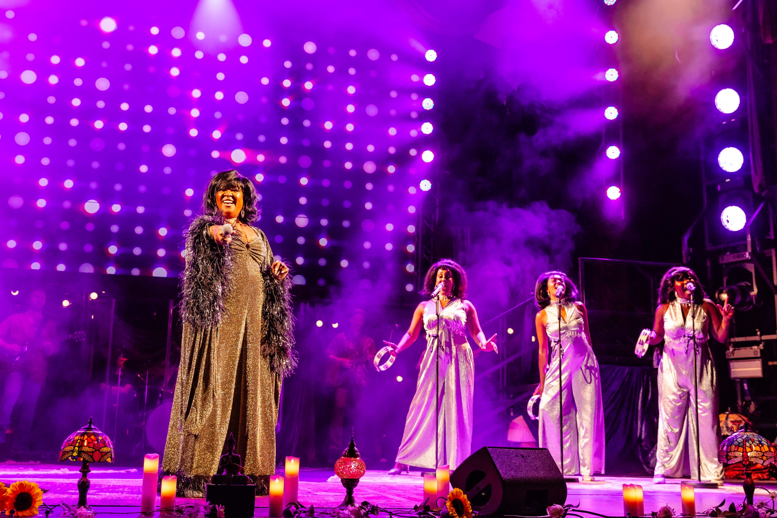 Still photo from the stage show A Night With Janis Joplin The Musical, on stage Kalisha Amaris wearing along gold dress singing infront of a microphone, with three backing singers behind her.