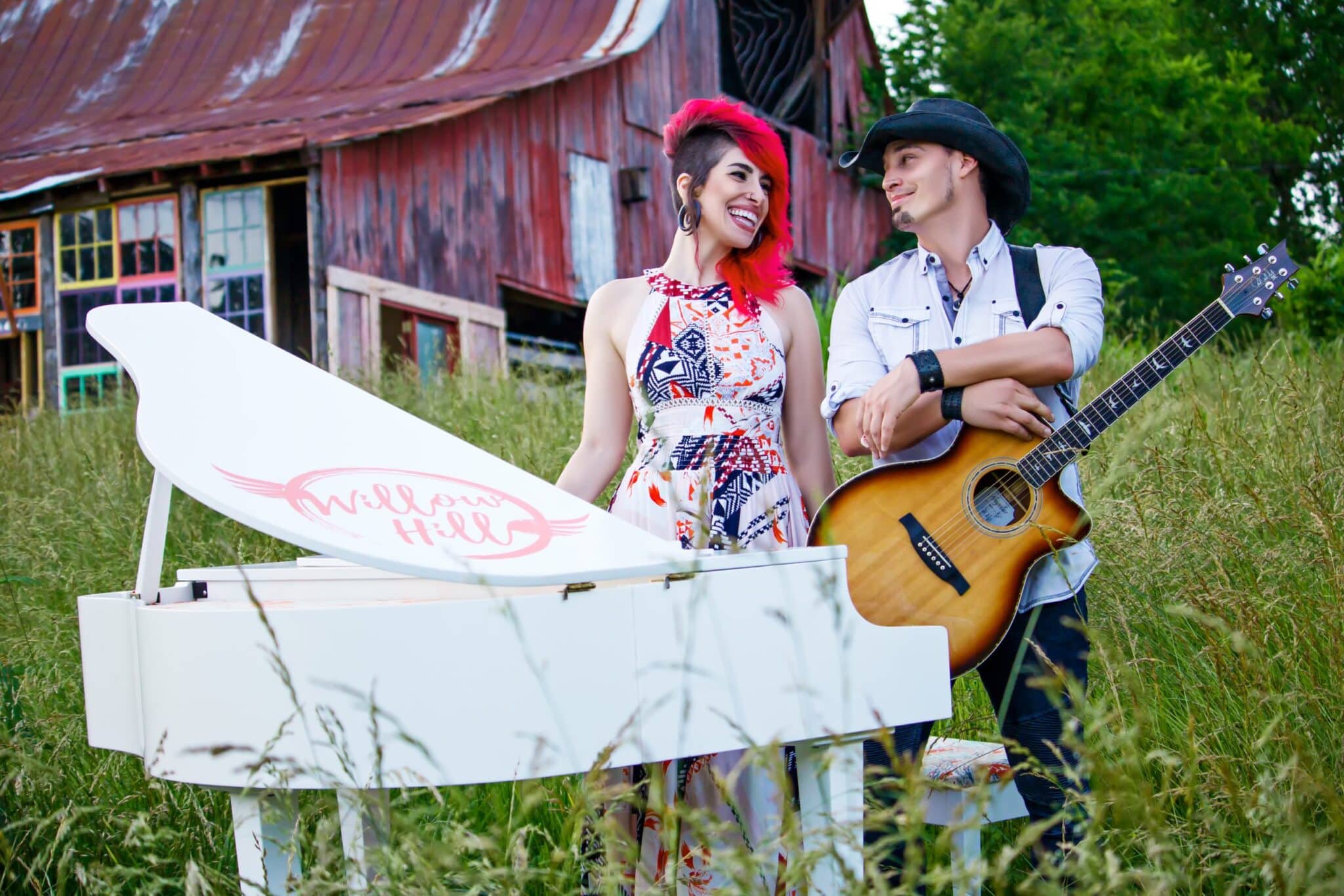 Willow Hill's Alexandra & CT standing behind a white grand piano that is amongst long grass, in front of a wooden barn. Alexandra has long dyed red hair and wearing a sleeveless floral dress, CT is wearing a white shirt and is carrying a guitar