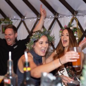 Mixo's Cocktail Master Class Party, two smiling ladies, one holding a cocktail, both wearing hair garlands, with Lee Cowell, Mixologist in the background, wearing black shirt, smiling and arms in the air