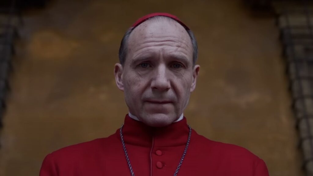 Ralph Fiennes as Cardinal Lawrence, wearing red vestments, looking towards the camera