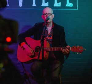 David Freja wearing a black jacket and playing his guitar singing into a microphone