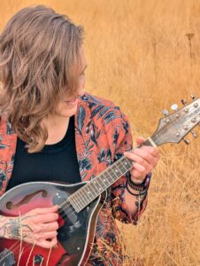 Kelly Wade, with long blonde hair, black vest and floral top playing a guitar