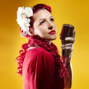 Miss Ruby Rouge vintage singer red dress dress and white flower in her hair holding a replica 1940s microphone