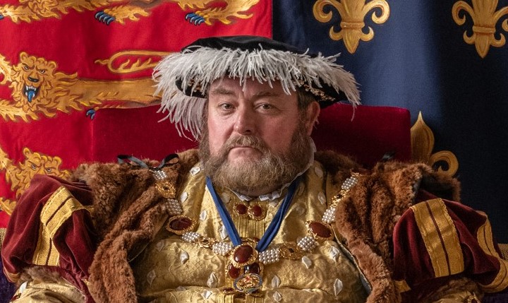 Jack Abbott dressed as King Henry 8th sat in front of a royal standard in Divorced, Beheaded, Died