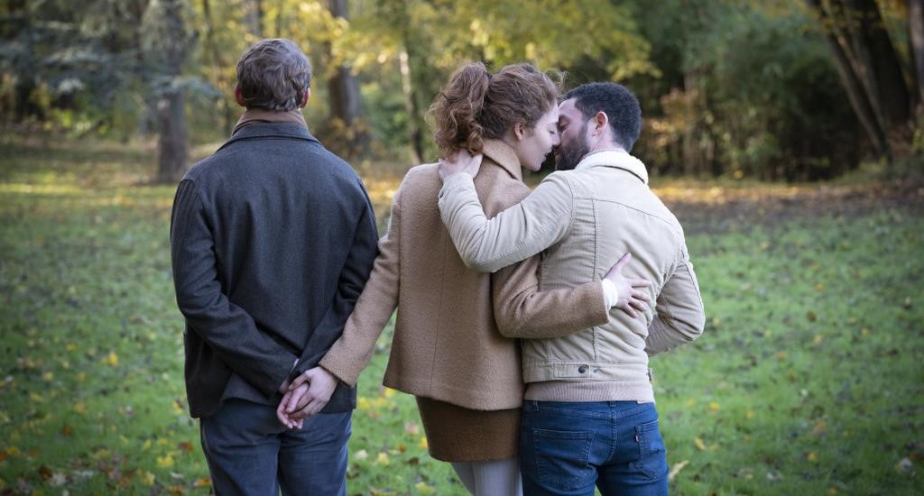 Rear view of Daphné (Camélia Jordana), Maxime (Niels Schneider) and François (Vincent Macaigne), walking on green grass towards some trees that are in leaf. Daphne is in the middle, wearing a brown jacket and black short skirt, she is holding hands with Maxime, is to her left, he is wearing blue jeans, dark grey jacket and light brown scraft, and looking straight head. Daphné’s head is turned to François, who is on her right, and she is kissing him. François is wearing blue jeans, and a sandy coloured sherpa jacket
