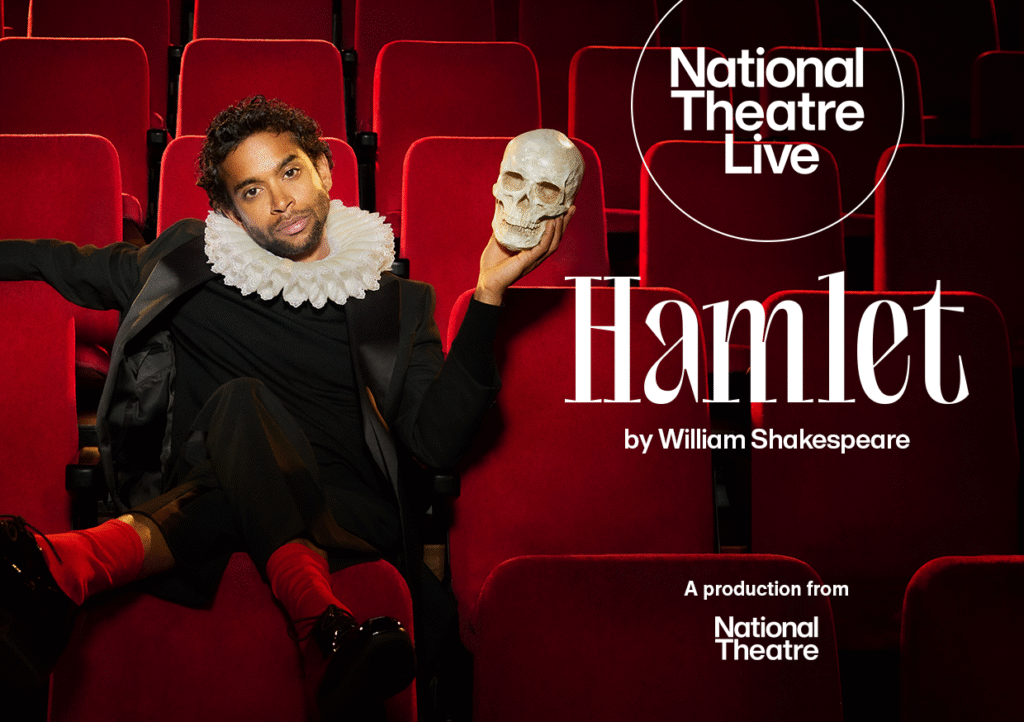 Hamlet played by Hiran Abeysekera sitting relaxed in a theatre seat, with legs hanging over the seat in front, wearing a long sleeved black top, black trousers, white ruff, red socks and held in his left hand a skull.