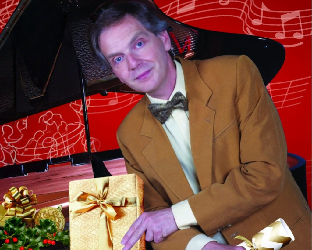 Michael Lunts wearing a white shirt, green bowtie and light brown suit jacket, leaning on a grand piano, his left hand holding a present wrapped in gold coloured paper and his left hand holding a gold coloured Christmas Cracker.