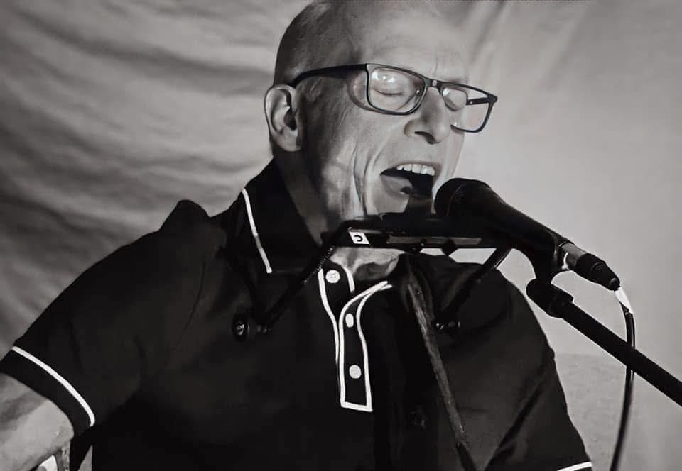 Paul Mills wearing black polo t-shirt with white piping, wearing glasses, singing in to a microphone, whilst playing a guitar
