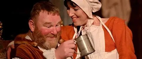 Falstaff played by Giles Shenton, in a cheery mode holding up a tankard, with Mistress Quickly, played by Georgie Taylor by his side