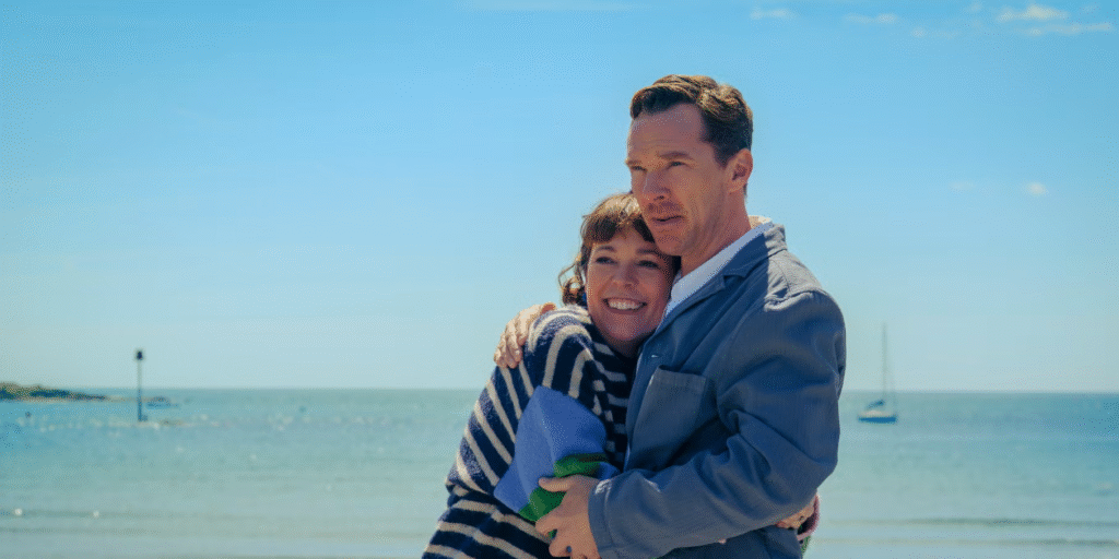 Standing on a beach with the sea behind them is Ivy (Olivia Colman) and Theo (Benedict Cumberbatch). Ivy is wearing a blue and white stripped top and Theo is wearing a blue jacket. Theo's arm is around Ivy's shoulder and her head is nestled under his chin.