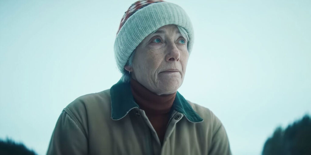 Emma Thompson wearing a red and white woolly hat, dark red jumper and an overcoat, looking thoughtfully with eyes looking up.