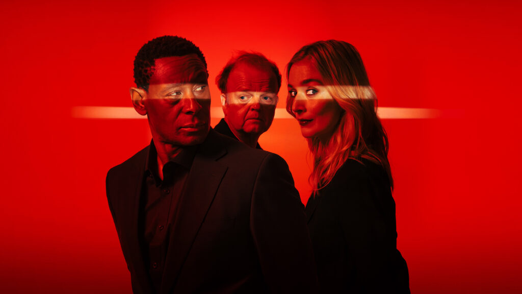 Above waist image for OTHELLO, David Harewood, Toby Jones and Caitlin Fitzgerald, each looking in a different direction, wearing black open neck suits, set against a red background