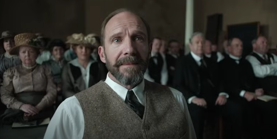Above waist photo of Dr. Henry Guthrie played by Ralph Fiennes looking forward, wearing white shirt, black tie, green-grey tweed waist cost, sporting a beard and moustache.
