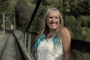 Vikki Clayton standing on a bridge, with trees in the background, wearing a white, sleeveless top. Vikki is smiling and has long blond hair.