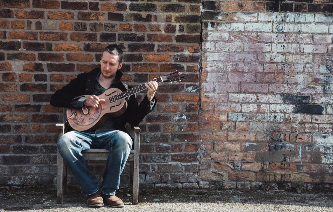 Gren Bartley wearing a black top and blue jeans, sitting on a wooden chair in front of a brick wall playing a guitar