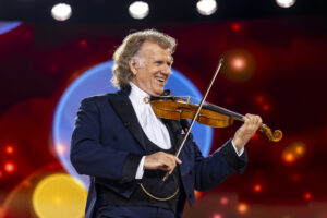 Andre Rieu Vrijthof - Bromsgrove Arts Smiling violinist in a dark suit playing a violin on a brightly lit stage with colorful background lights.