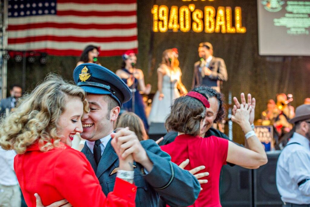 Couple dancing closely in vintage 1940s attire at a ball, with a live band on stage and a large '1940's Ball' sign in the background.