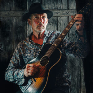 Photos of Paul Cowley, wearing a blue shirt, black cowboy hat, playing a guitar, taken by Karen Cooper