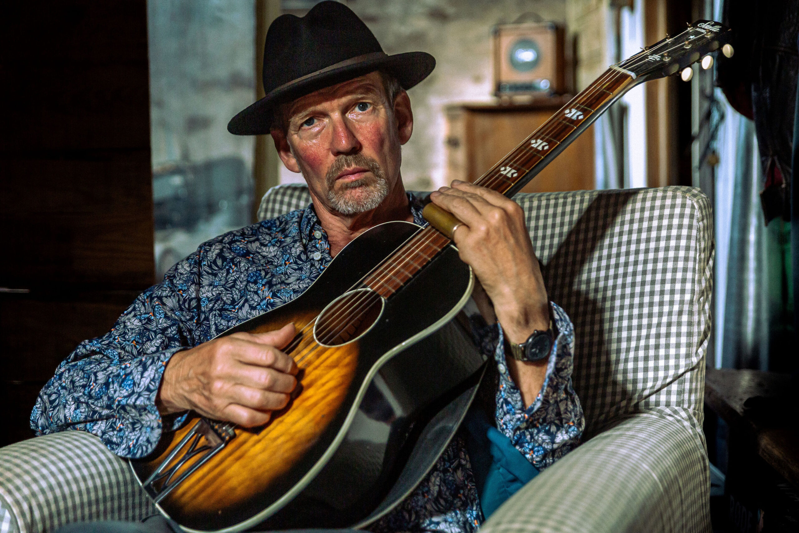 Photos of Paul Cowley, wearing a blue shirt, black cowboy hat and playing a guitar, whilst sat in a grey arm chair, taken by Karen Cooper