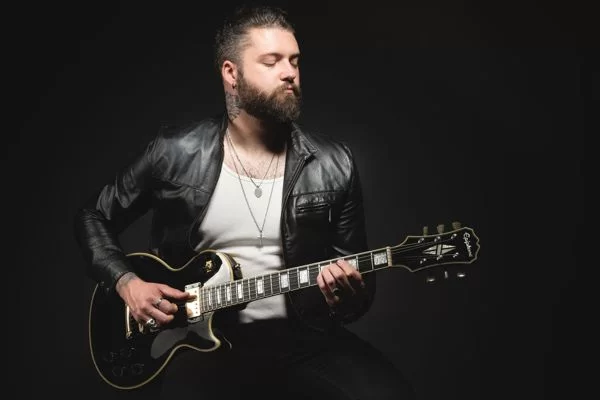 Ross Harding with beard, wearing black leather jacket over a white t-shirt. Ross is playing an electric guitar