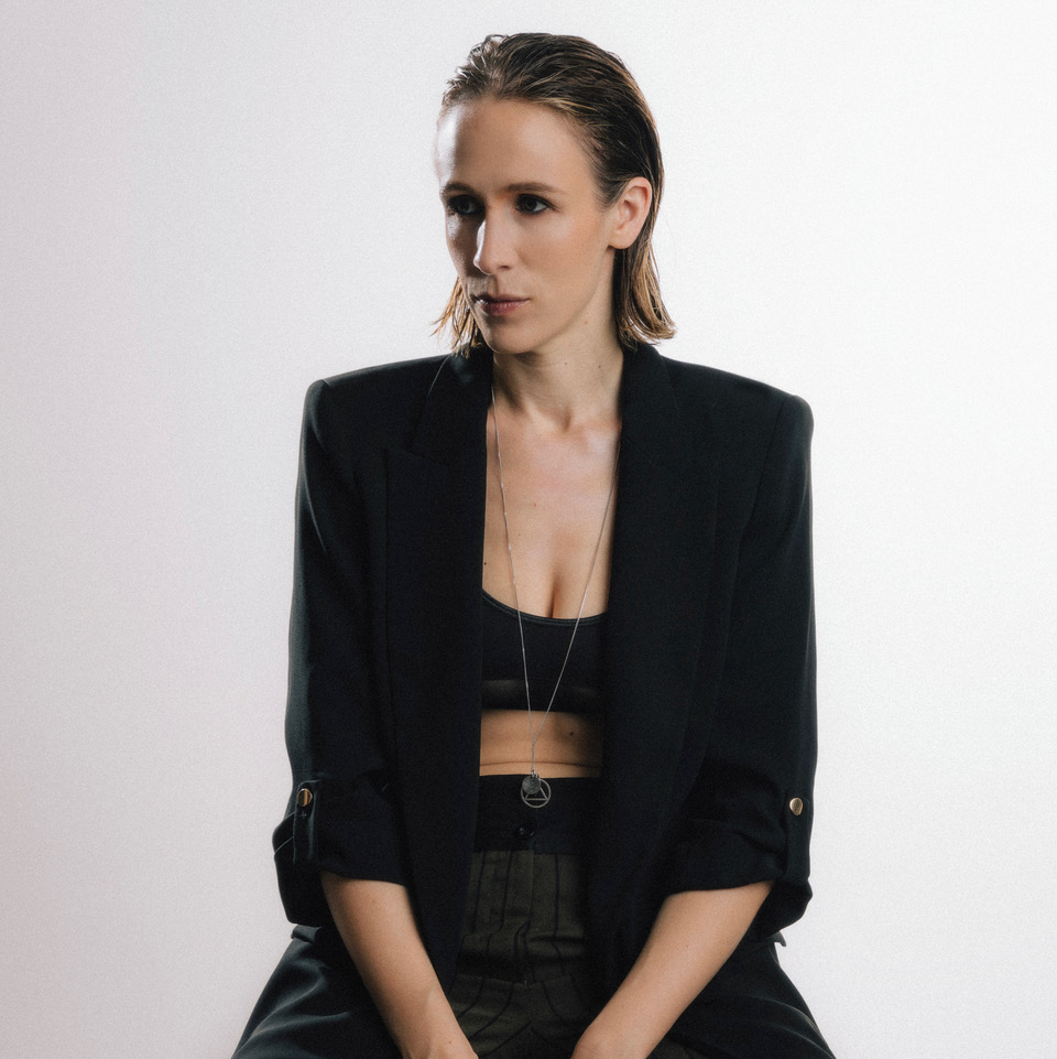 Portrait of a Katey Brooks in a black blazer and crop top, wearing a long silver pendant necklace, looking to the side against a light background.