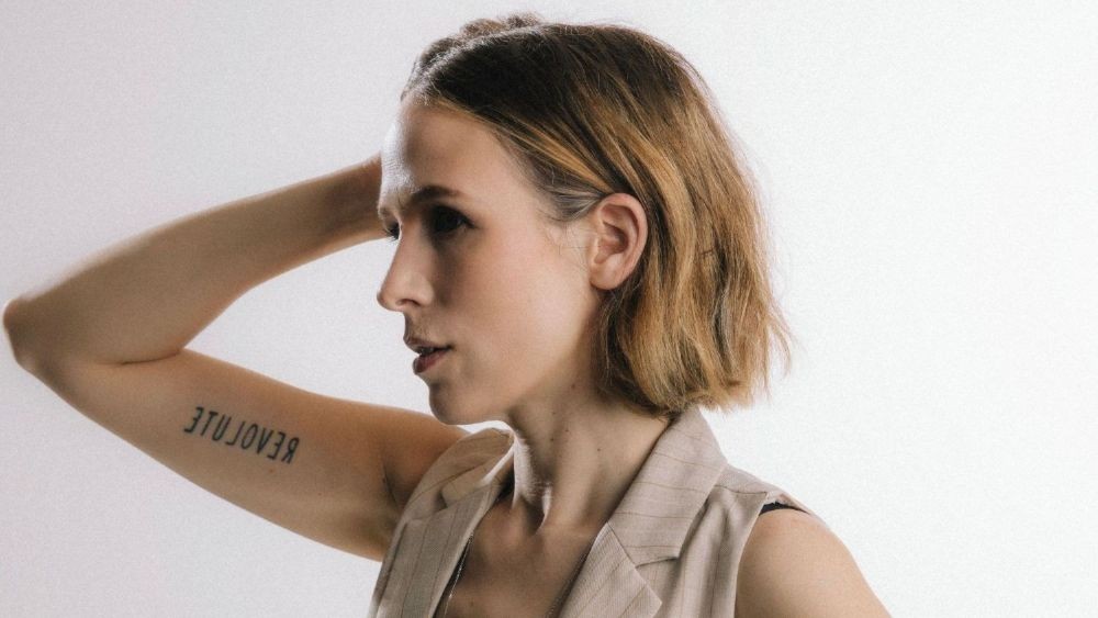 Side profile of a Katey Brooks with short, light brown hair wearing a beige sleeveless blazer, arm raised behind her head, exposing a tattoo on her inner forearm.