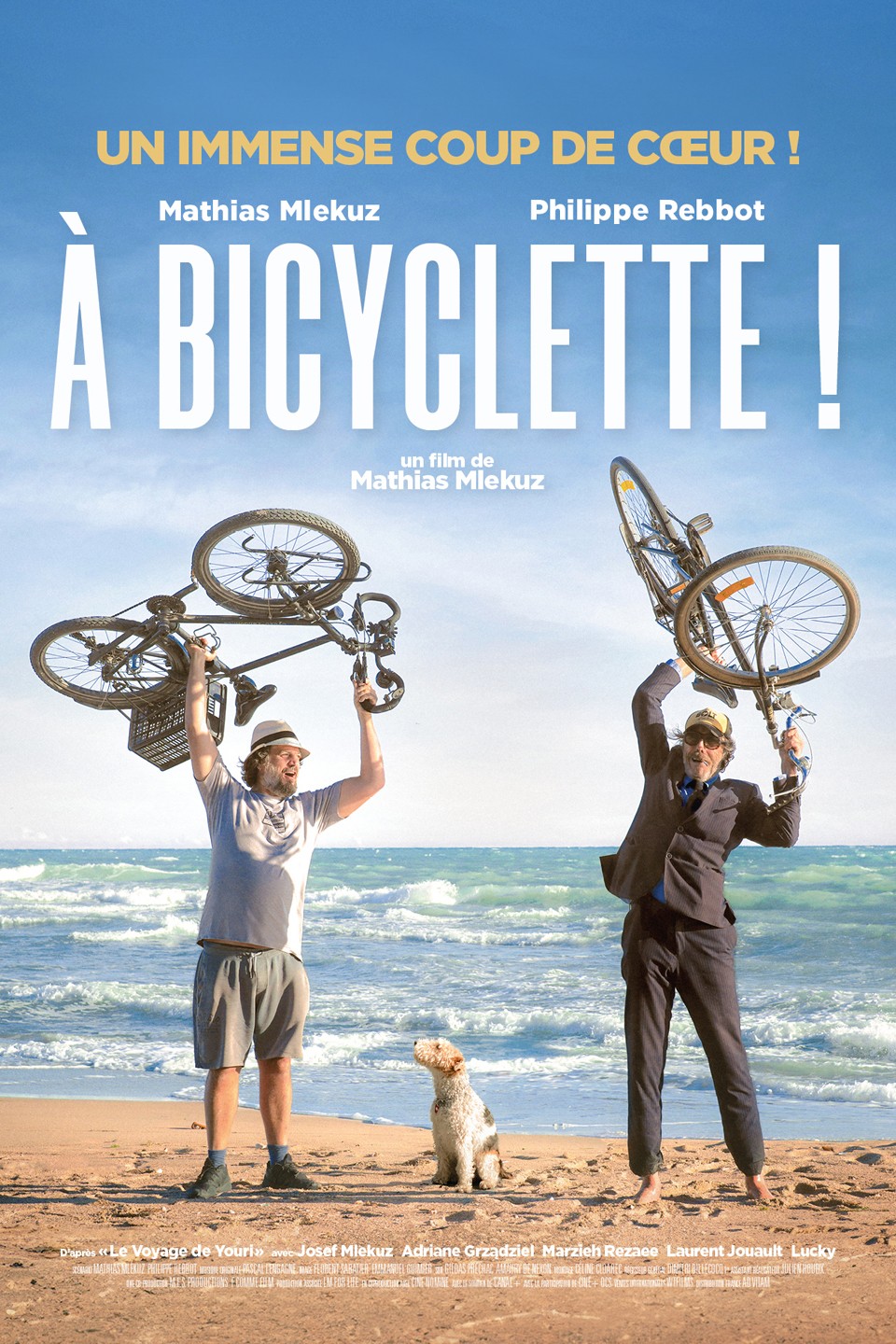 Mathias Mlekuz & Philippe Rebbot standing on a sandy beach, the sea behind them, a dog sat between them, and both men holding their bicycles above their heads