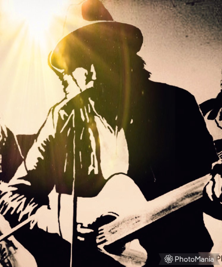 Tom Martin, wearing wide brimmed hat, sunlight above, playing a guitar and singing into a microphone, which is on a stand in front of him.