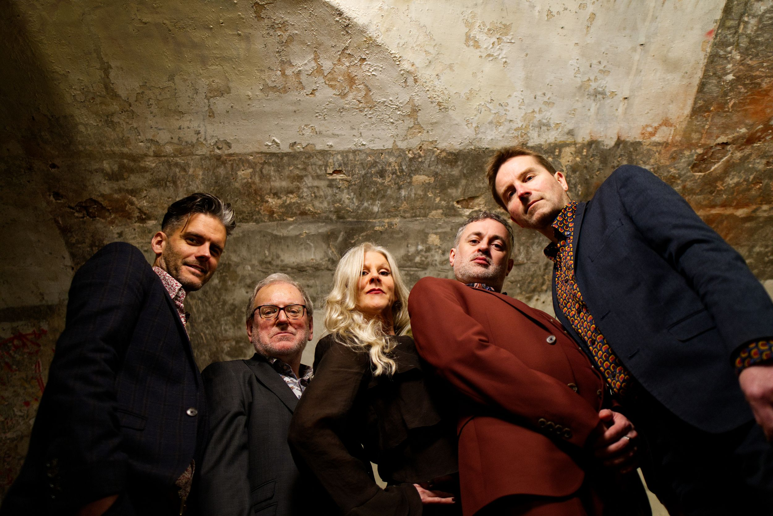 MDC Ceiling Low Resjpg - Bromsgrove Arts Six people in formal attire lean toward the camera from a low angle in a worn, concrete space with peeling walls.