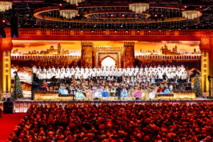 Large choir in white robes plus an orchestra performing on an ornate-lit stage with a mural of ancient city walls and arches in the background.