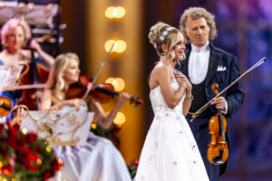 Smiling bride in a white wedding dress with a snowflake headpiece on stage beside an orchestra of musicians in gowns and suits.
