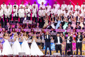 Large orchestra and choir perform on a brightly lit stage, with men in white tuxedos and women in white gowns in the foreground and background.