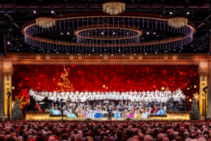 Large festive choir on a decorated stage with an orchestra and red holiday backdrop, audience in the foreground.