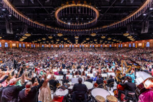 Wide view of a full orchestra performing on stage in a grand concert hall, with the conductor center and a packed audience beyond.