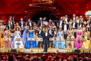 Conductor leads a full orchestra and choir on a decorated stage with a red festive backdrop and audience visible in front.