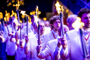 A group of people in white outfits marching with lit torches at night, illuminated by colorful purple and blue lights.