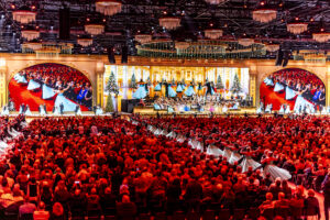 Large concert hall with dancers in turquoise gowns performing on a grand stage, audience in red seats, festive decorations aritistically lit.