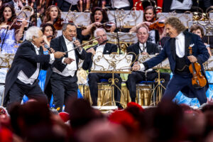 Three tuxedo-clad performers sing and gesture on a concert stage with a full orchestra behind them, audience in the foreground.