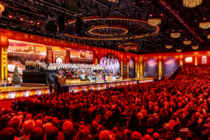 Full orchestra and choir performing on a festive-lit stage in a grand concert hall, with red ambience and holiday decor.