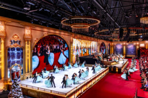 Grand ornate theater stage with dancers in turquoise gowns performing before a gold-decorated backdrop and a large audience