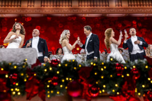 Group of formally dressed performers on a stage toasting champagne against a red festive backdrop.