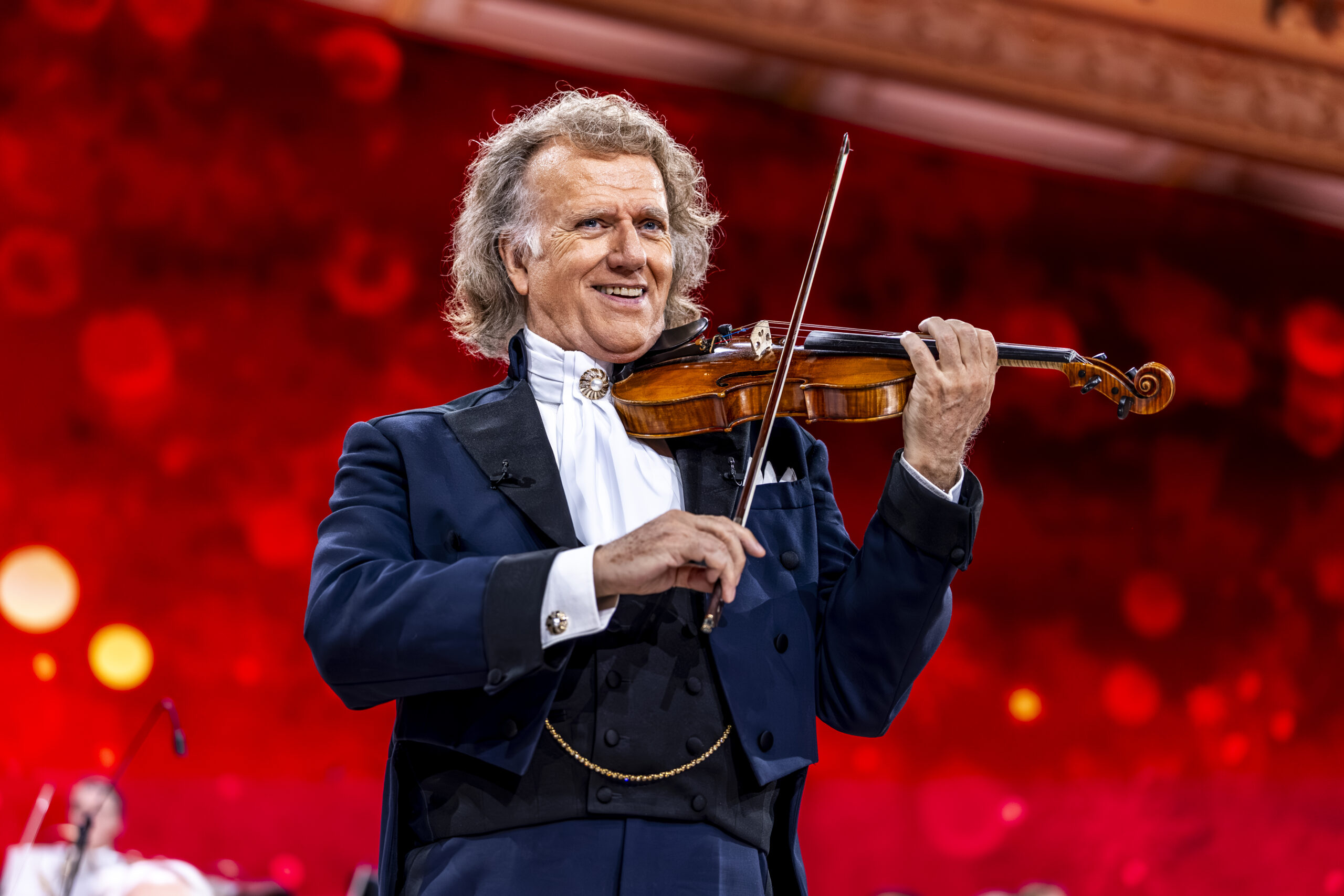 In front of a red back ground, Andre Rieu playing his violin, wearing his trademark blue jacket, white shirt, the collar held together by a broach.