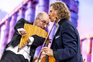 Two male musicians in tuxedos perform on a colorful stage; one plays a vertical pan flute while the other leans in close, holding a violin.