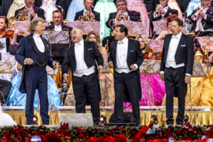 Four men in tuxedos singing on a decorated concert stage, with an orchestra in the background and colorful dresses visible behind them.