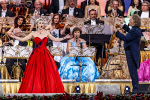 Soprano in a red ball gown sings with a full orchestra on a decorated stage.